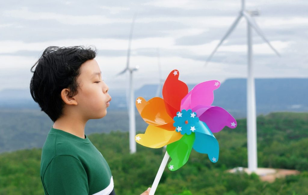 Progressive young asian boy playing with wind pinwheel toy in the wind turbine farm, green field over the hill. Green energy from renewable electric wind generator. Windmill in the countryside concept