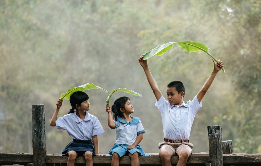 Asian boy and little girls sitting on wooden bridge and joyful to playing with banana leaves on head, smile and laughting with funny together, copy space, rural scene style concept